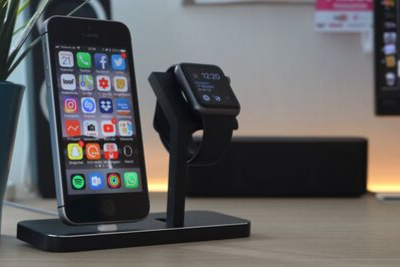 A black apple watch without a protector case, sits on a wireless charging station next to an iPhone.