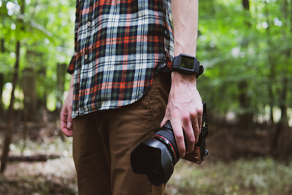 Man with camera in a plaid shirt and khaki pants, stands in a forest wearing Rhino Brand's black Apple Watch Protector.